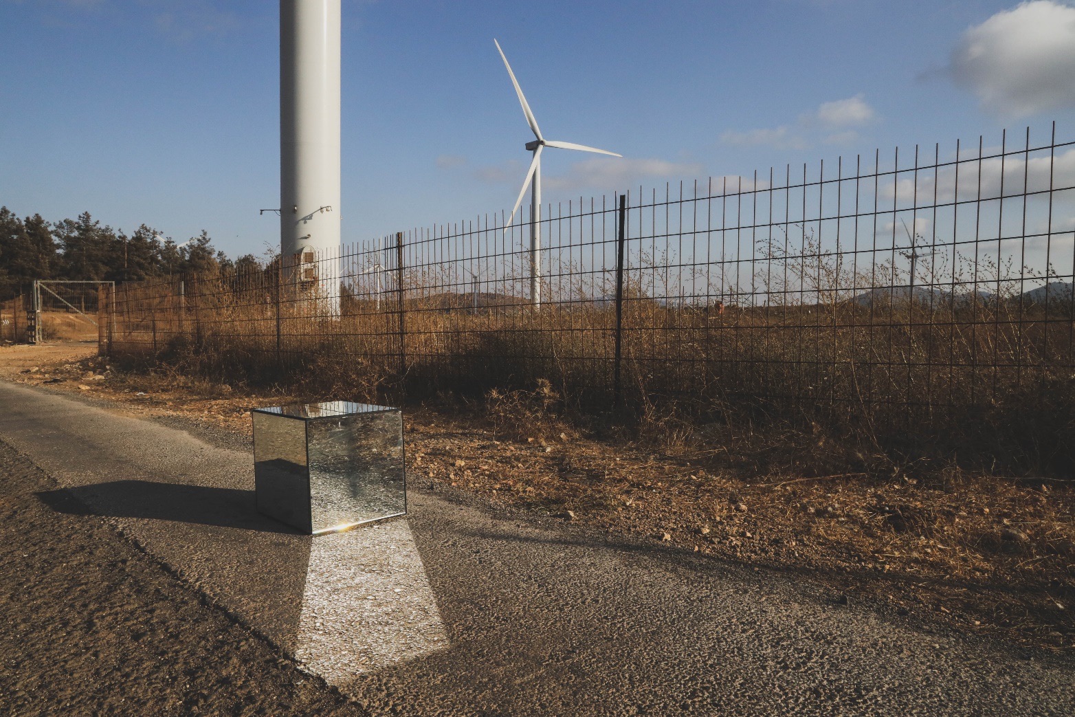 A mirrored cube on a road beside a fenced wind turbine field